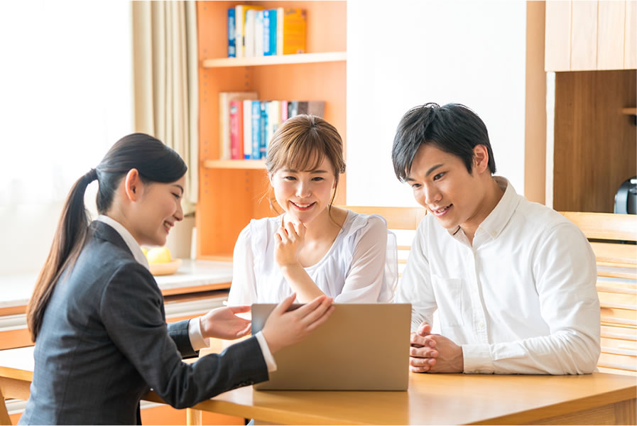 alt="住宅会社の担当者が夫婦にパソコンを使って土地や建物、資金計画について説明している様子。安心感のある打ち合わせ風景。"