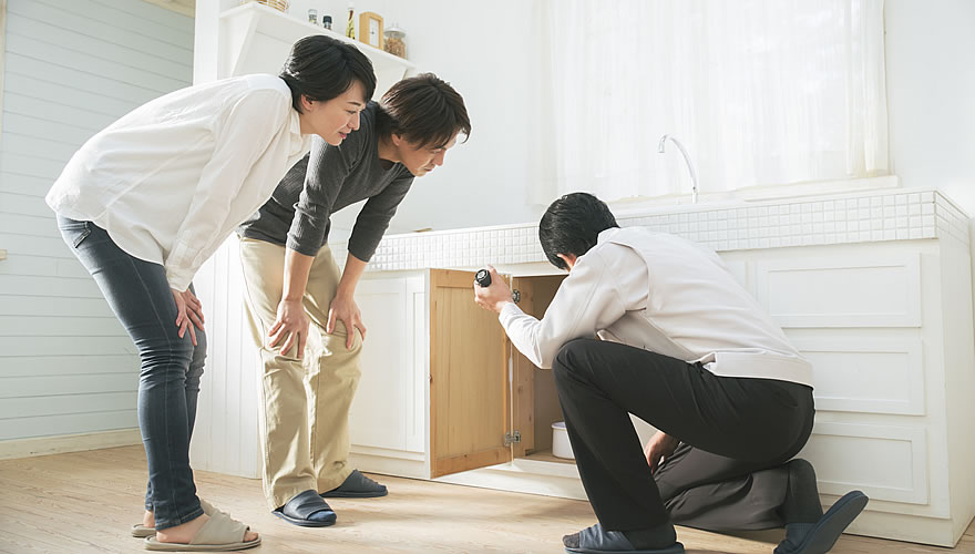 alt="住宅のアフター点検で、スタッフがシンク下の収納を点検しながら施主に説明している様子。地域密着の丁寧な対応をイメージ。"