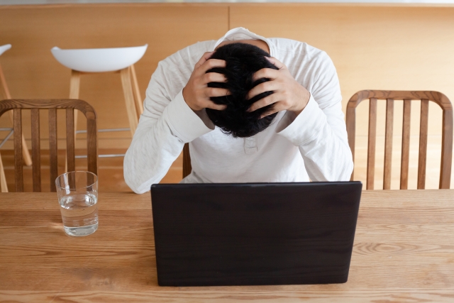 alt属性="ノートパソコンの前で頭を抱える男性|家づくりの後悔や不安をイメージ"