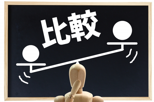 alt="比較を表す黒板イラスト。シーソーに乗った2つの円が描かれ、中央で木製人形が考えている様子が示されている。標準仕様とオプションの違いを象徴するイメージ。"
